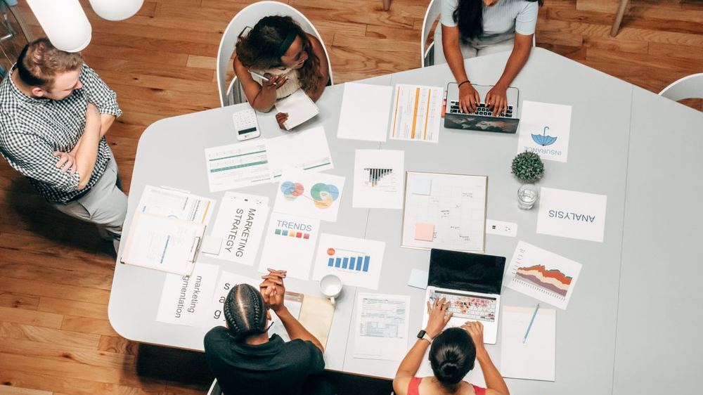 Team aan een vergadertafel vol uitgeprinte plannen en laptops, van bovenaf gezien